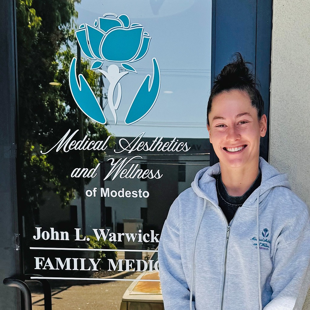 Professional athlete Nicole Warwick smiling in front of the Medical Aesthetics & Wellness of Modesto office wearing an MAW hoodie, representing the partnership promoting full-body health and wellness.